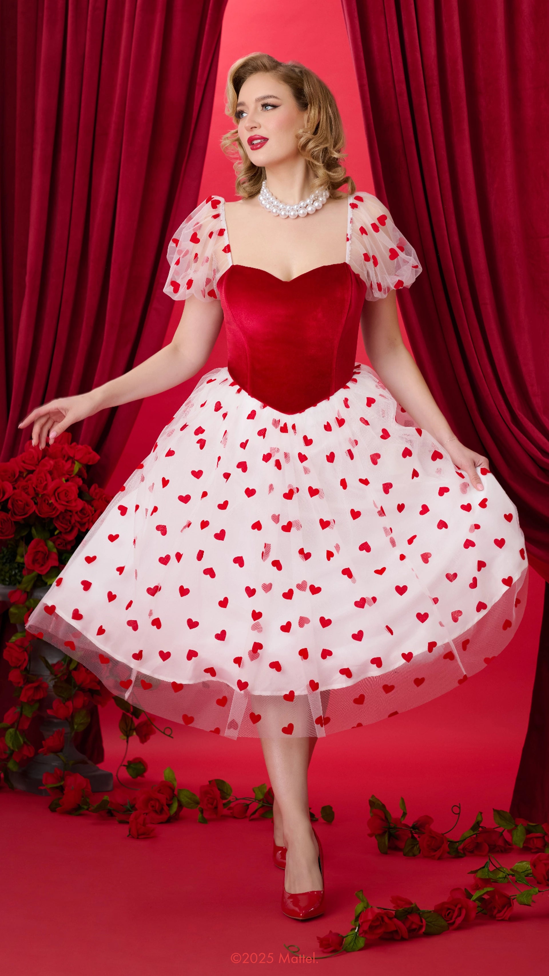 Woman wearing a red and white dress with heart patterns against a red curtain background.