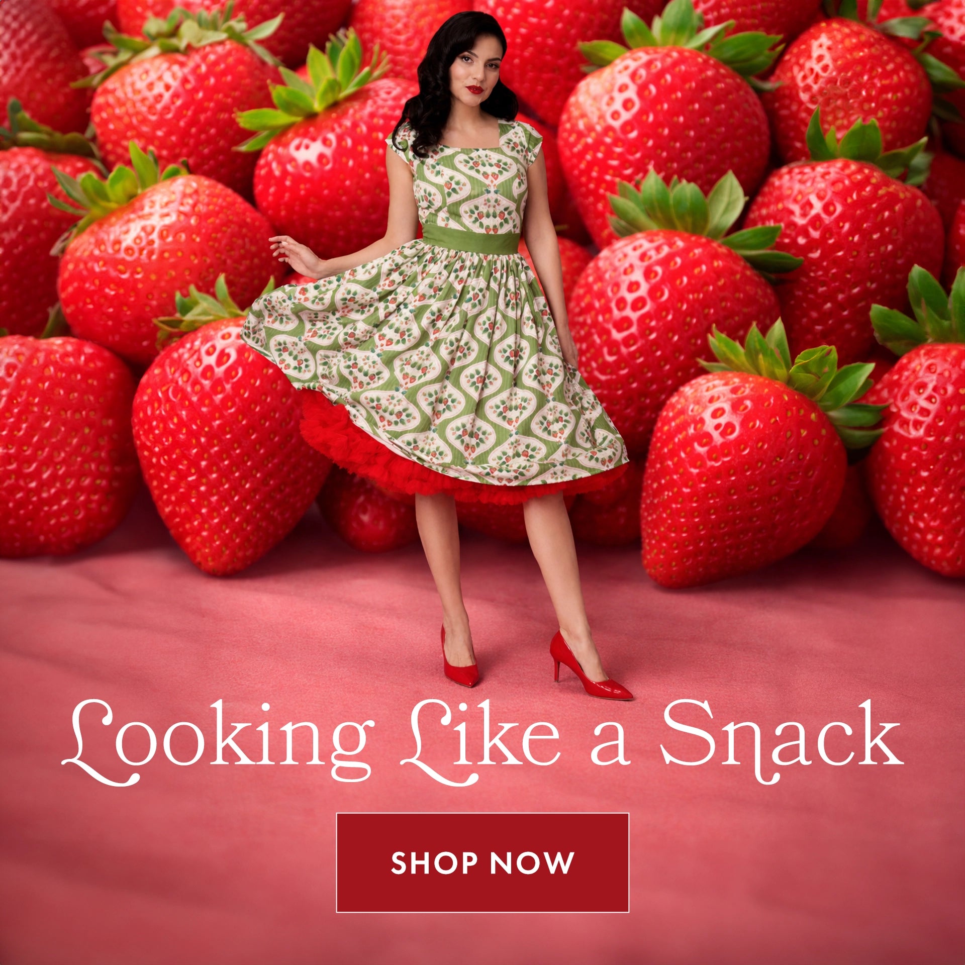 A woman wearing a vintage-style green and cream patterned dress with a red petticoat and red high heels poses in front of an oversized background of bright red strawberries. She is mid-twirl, holding the skirt out slightly. At the bottom, stylized text reads “Looking Like a Snack” with a red “Shop Now” button below.