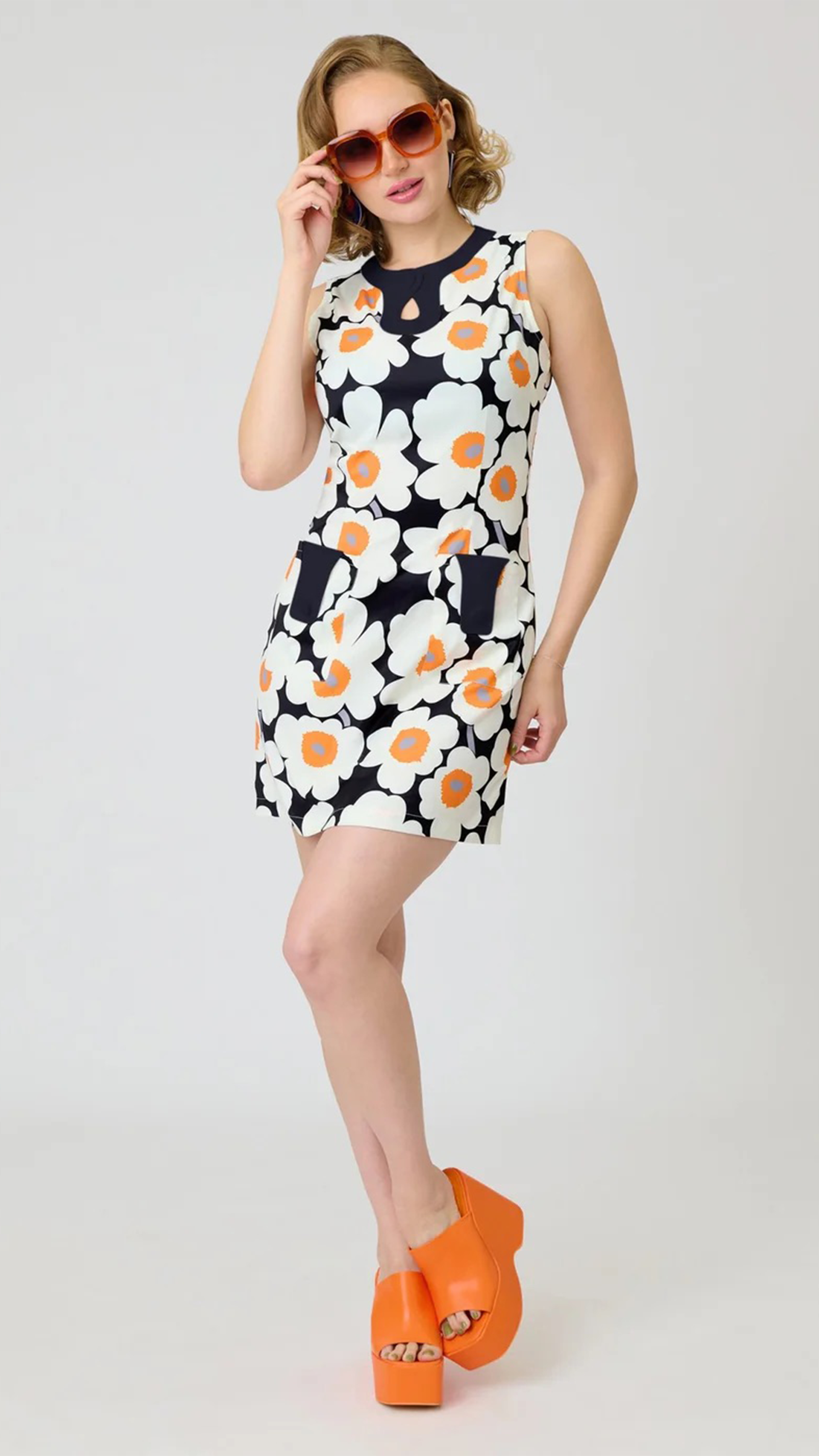 A woman modeling a sleeveless mini dress with a bold white, orange, and navy floral pattern, styled with orange platform sandals and orange-tinted sunglasses against a light gray studio background.