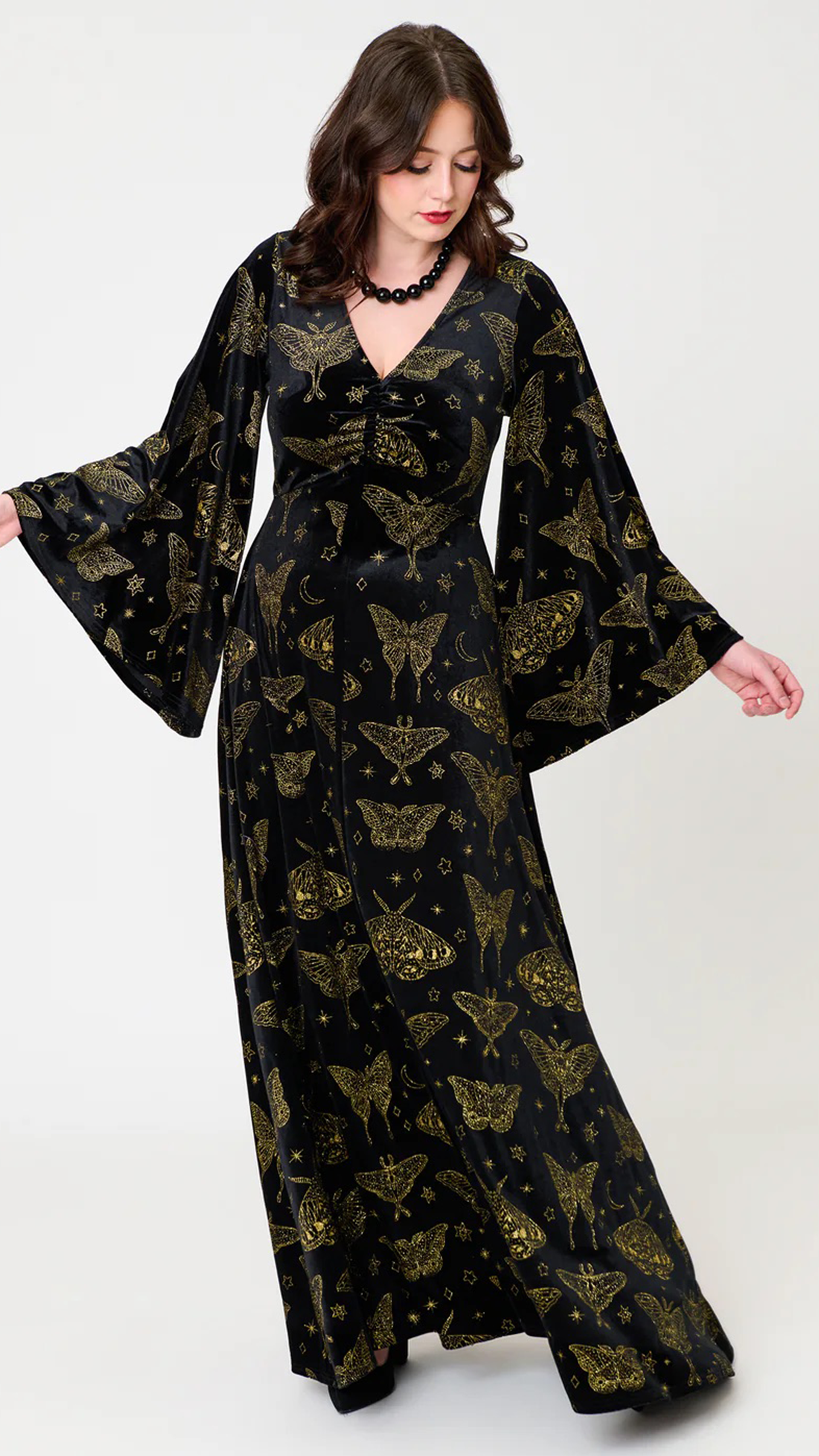 A woman modeling a long black maxi dress with a gold metallic butterfly and star pattern, featuring wide bell sleeves and a V-neckline, set against a plain light gray studio background.