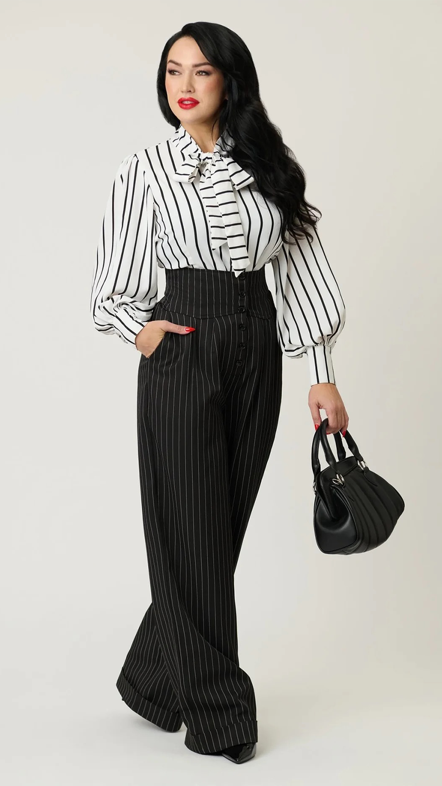 A woman modeling a black and white vertically striped blouse with puffed sleeves and a bow collar, paired with matching black pinstriped wide-leg trousers, holding a black quilted handbag against a light gray studio background.