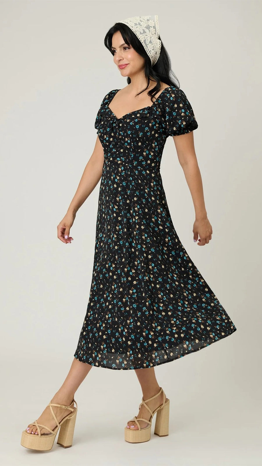 A woman modeling a black midi dress with a small multicolor floral print, featuring puff sleeves and a square neckline. She is wearing a white lace headband and beige platform sandals against a plain light gray studio background.
