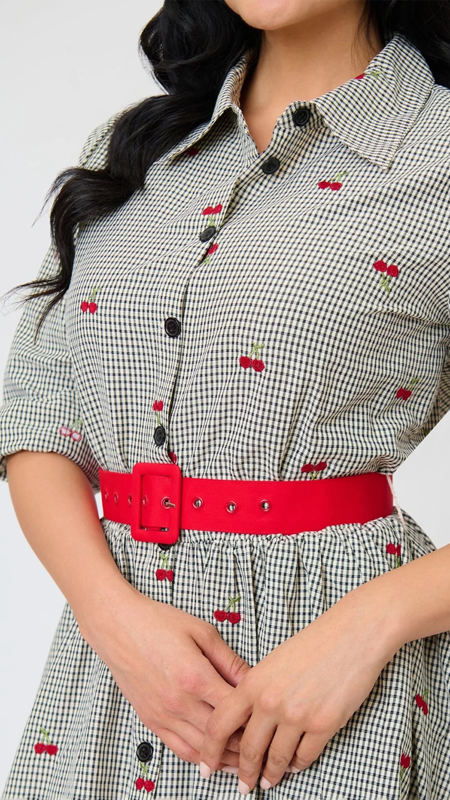 A close-up of a person wearing a black and white gingham shirt dress with small red cherry prints, a bright red belt with a silver buckle, and black buttons.