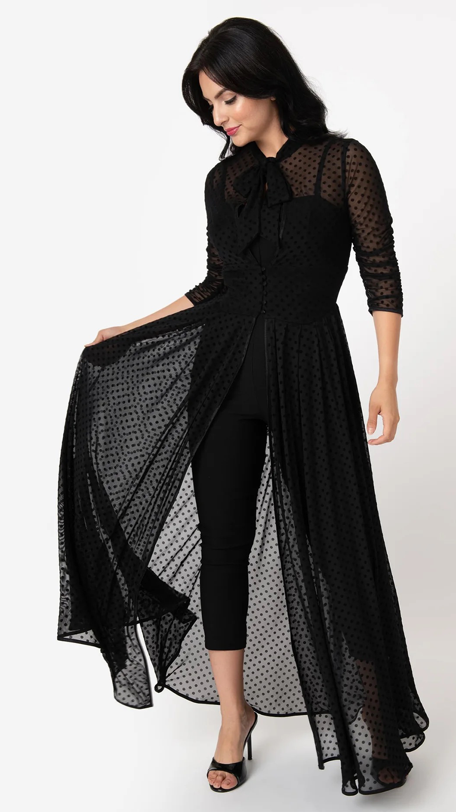 A woman modeling a black sheer long-sleeve top with a polka dot pattern, a high-low draped overlay skirt, and a tie-neck bow, paired with black cropped pants and black heeled sandals against a white studio background.