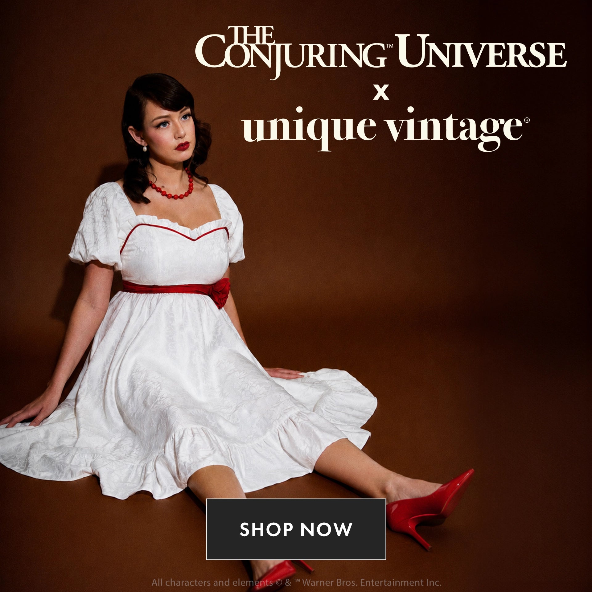 A woman seated on a brown studio background wearing a white vintage-style dress with red trim and a red sash, red heels, and a red necklace. Large text at the top reads 'The Conjuring Universe x unique vintage' and a 'SHOP NOW' button appears at the bottom.