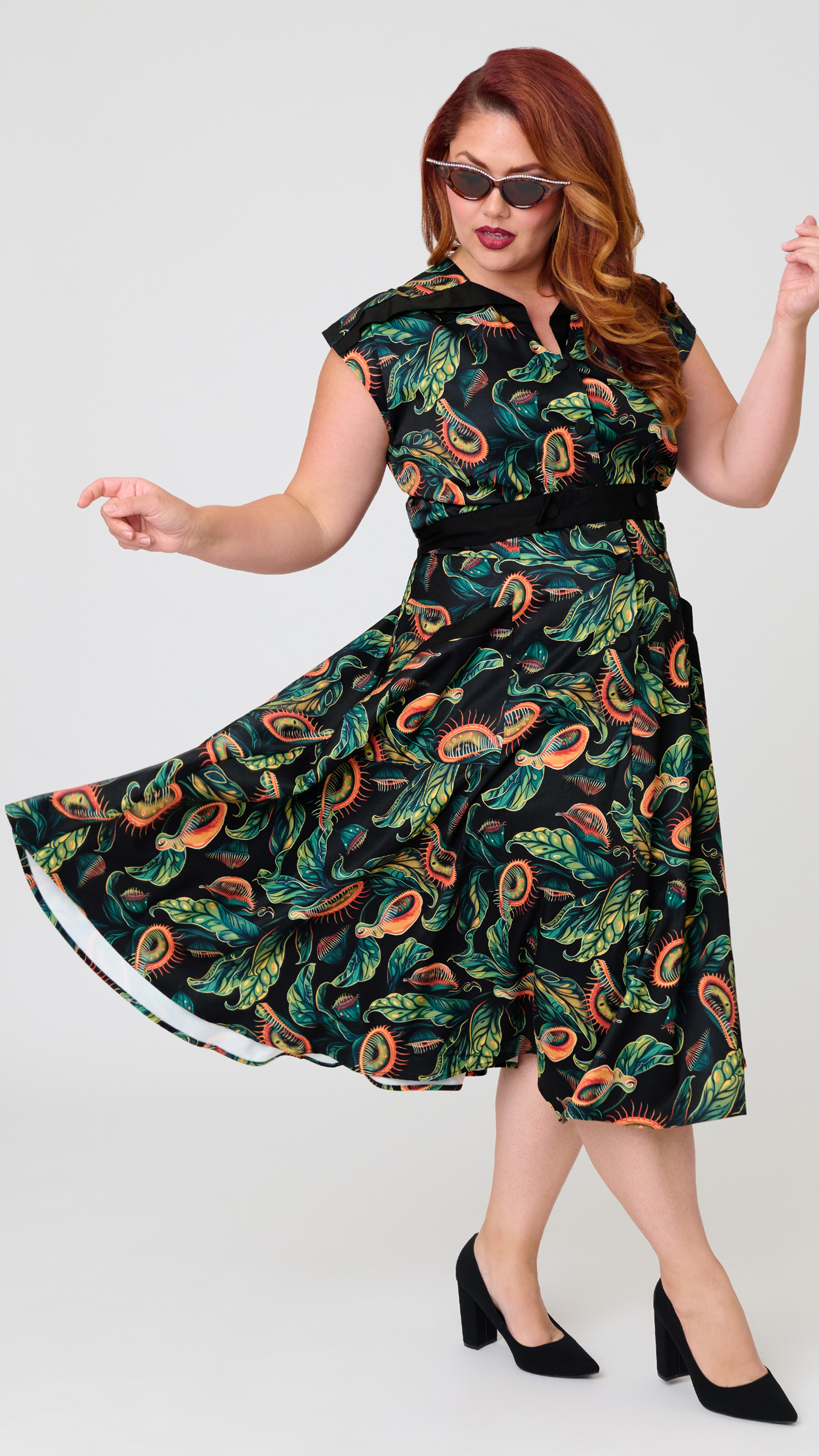 A woman modeling a black midi dress with a colorful retro paisley and leaf pattern in green, orange, and yellow, paired with black high heels against a light gray studio background.